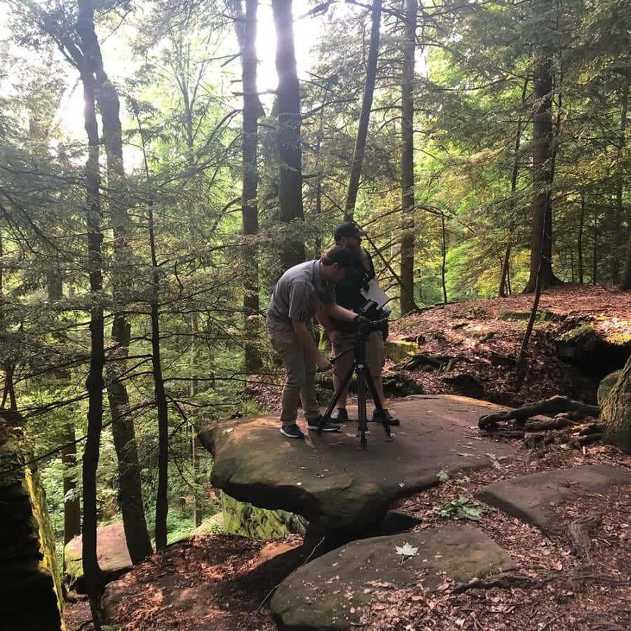 Two people running a camera on a tripod, standing on a large flat rock in a forrest